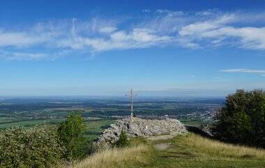 Eine hügelige Landschaft mit einem kleinen Gipfelkreuz und weitem Blick über Felder und einen klaren blauen Himmel.
