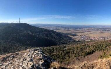 Die Panoramaaufnahme zeigt eine hügelige Landschaft mit einem bewaldeten Gebirge im Vordergrund und einer weiten Ebene im Hintergrund unter einem klaren blauen Himmel.